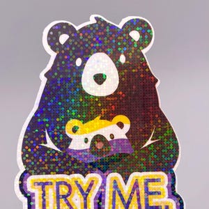 May include: Holographic sticker featuring a bear holding a smaller bear, with the text "TRY ME" in yellow and purple. The bears have a dark brown body with a shimmering, multi-colored effect.