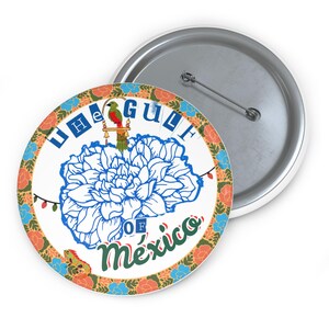 Könnte beinhalten: Ein weißer Knopf mit einem bunten Rand, der eine blaue Blume und den Text "The Gulf of Mexico" zeigt. Ein kleiner Vogel sitzt auf einem Ast über der Blume.