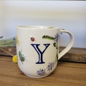 May include: A white ceramic mug featuring a large, dark blue letter Y in the center. The mug is decorated with raised, colorful illustrations of a ladybug, a green caterpillar, a green beetle, and light blue borage flowers around the letter.