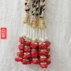 May include: A decorative hanging ornament featuring three black braided cords adorned with gold metallic cone-shaped tassels and white pearl beads. The bottom section consists of multiple layers of red fabric rose petals with gold trim.