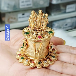 May include: A gold-coloured decorative item featuring three figures. It is embellished with red and green gemstones and detailed patterns. The text "Design -1" is visible on the left side.