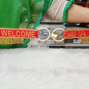 May include: A red fabric banner with gold trim and fringe featuring the word WELCOME in gold lettering. Two white pearl-covered rings are linked together in the center, secured by a small brass padlock with a visible keyhole on the front.