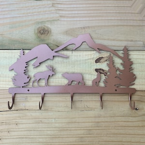May include: A copper-colored metal wall hook rack featuring a laser-cut silhouette of a mountain range, pine trees, a moose, a bear, and a howling wolf. The functional piece includes five downward-curving hooks along the bottom edge.