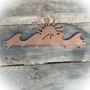 May include: A copper-colored metal key rack shaped like a sun setting over waves. The rack has five hooks.