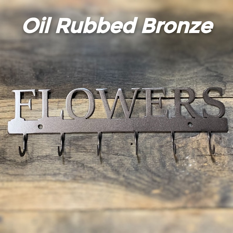 Puede incluir: Perchero de pared de metal bronce al aceite con la palabra "FLOWERS" recortada. El gancho tiene seis ganchos curvos debajo de la palabra. El fondo es una superficie de madera.