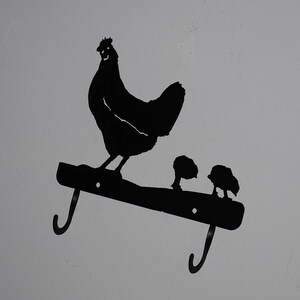May include: Black metal silhouette of a hen and two chicks on a hook rack. The hen is standing on a branch with her chicks below her. The hook rack has two hooks.