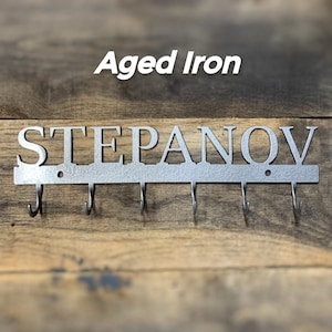 Puede incluir: Llavero de pared de hierro envejecido con el nombre "STEPANOV" recortado en may&uacute;sculas. El llavero tiene cinco ganchos y est&aacute; montado sobre un fondo de madera. El texto "Aged Iron" est&aacute; encima del nombre.
