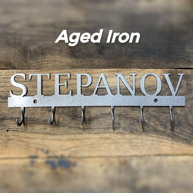 Puede incluir: Llavero de pared de hierro envejecido con el nombre "STEPANOV" recortado en may&uacute;sculas. El llavero tiene cinco ganchos y est&aacute; montado sobre un fondo de madera. El texto "Aged Iron" est&aacute; encima del nombre.
