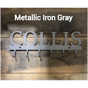 Puede incluir: Llavero met&aacute;lico gris hierro metalizado con el nombre "COLLIS" recortado. El llavero tiene cinco ganchos debajo del nombre y est&aacute; montado sobre un fondo de madera.