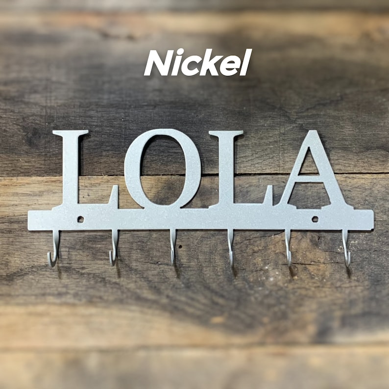 Puede incluir: Un llavero de pared de metal color n&iacute;quel con la palabra "LOLA" y cinco ganchos. La palabra "Nickel" est&aacute; encima del nombre. El llavero est&aacute; montado sobre un fondo de madera.