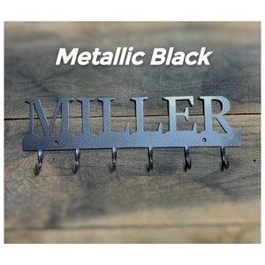 Puede incluir: Llavero de metal negro metalizado con el nombre "MILLER" recortado. El llavero tiene seis ganchos para llaves y est&aacute; montado sobre un fondo de madera. El texto "Metallic Black" est&aacute; encima del nombre.