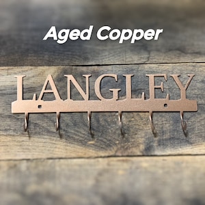 Puede incluir: Llavero de metal de cobre envejecido con el nombre "LANGLEY" recortado. El llavero tiene cinco ganchos y est&aacute; montado sobre un fondo de madera. El texto "Aged Copper" est&aacute; encima del nombre.