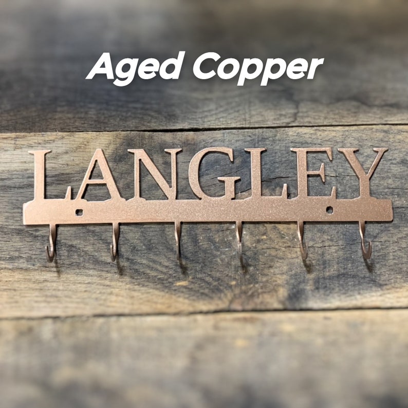 Puede incluir: Llavero de metal de cobre envejecido con el nombre "LANGLEY" recortado. El llavero tiene cinco ganchos y est&aacute; montado sobre un fondo de madera. El texto "Aged Copper" est&aacute; encima del nombre.