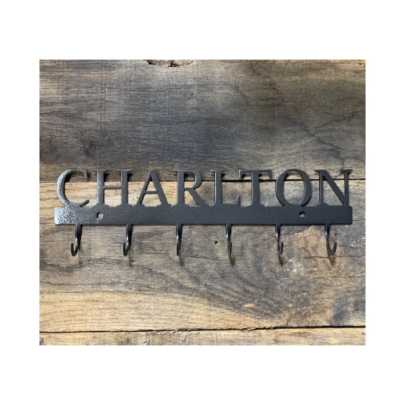 May include: Black metal key holder with the name "CHARLTON" cut out in capital letters. The key holder has six hooks below the name. The key holder is mounted on a rustic wooden background.