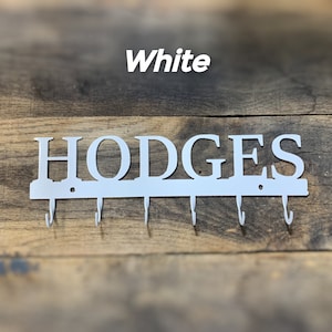 Puede incluir: Llavero de metal blanco con el nombre "HODGES" recortado. El llavero tiene seis ganchos debajo del nombre. La palabra "White" est&aacute; encima del nombre. El llavero est&aacute; montado sobre una superficie de madera.