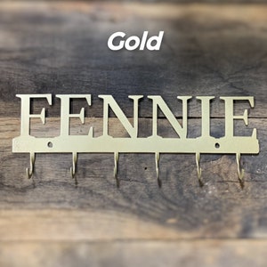 Puede incluir: Llavero de pared de metal dorado con el nombre "FENNIE" recortado. El llavero tiene seis ganchos debajo del nombre. La palabra "Gold" est&aacute; escrita encima del nombre en blanco.