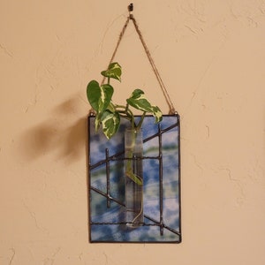 May include: A hanging stained glass propagation station with a clear glass tube holding a green and white plant. The glass has a blue and green watercolor design with black metal grid lines. A natural rope is used for hanging.
