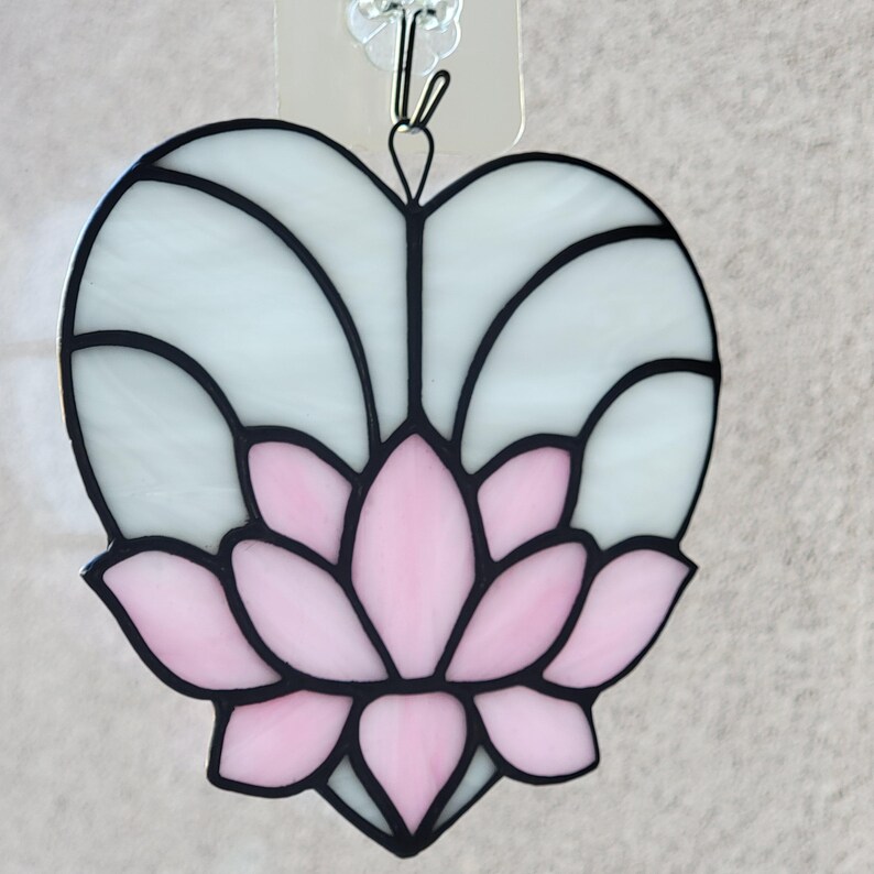 May include: Stained glass heart-shaped suncatcher featuring a pink lotus flower design. The lotus is set against a white background with black outlines. A metal hook is attached for hanging.
