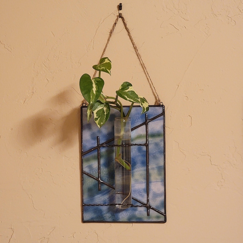 May include: A hanging stained glass propagation station with a clear glass tube holding a green and white plant. The glass has a blue and green watercolor design with black metal grid lines. A natural rope is used for hanging.