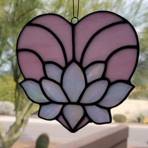 May include: A stained glass heart-shaped suncatcher with a lotus flower design. The heart is light pink with black outlines, and the flower is white with iridescent accents. A black wire loop is at the top for hanging.