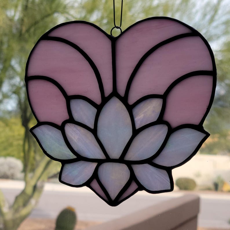 May include: A stained glass heart-shaped suncatcher with a lotus flower design. The heart is light pink with black outlines, and the flower is white with iridescent accents. A black wire loop is at the top for hanging.