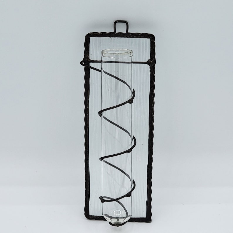 Peut inclure: Un cadre en m&eacute;tal noir avec un tube en verre transparent &agrave; l'int&eacute;rieur. Le tube est maintenu en place par un fil noir qui s'enroule autour de lui. Le cadre est mont&eacute; sur un mur blanc.