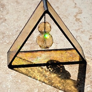 May include: A small triangular stained glass prism with a gold iridescent finish. A crystal prism hangs from the top of the prism and is suspended by a chain.