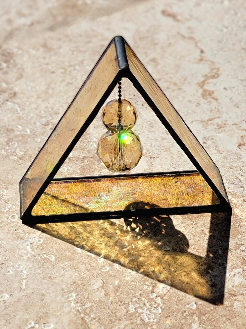May include: A small triangular stained glass prism with a gold iridescent finish. A crystal prism hangs from the top of the prism and is suspended by a chain.