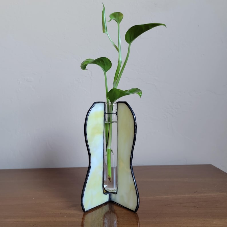 May include: A yellow stained glass vase with a clear glass tube in the center. A green plant is in the tube. The vase is on a wooden surface.