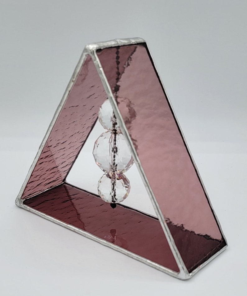 May include: A small triangular stained glass sculpture with a pink iridescent finish. Three clear crystal beads hang from the top of the triangle.