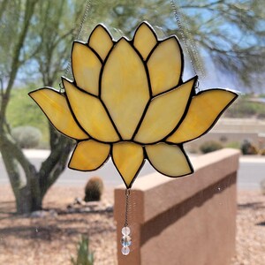 Peut inclure: Un attrape-soleil en forme de fleur de lotus en vitrail aux tons jaune et ambre. La fleur a des contours noirs et est suspendue par une cha&icirc;ne. Une petite cha&icirc;ne de perles pend du bas. L'arri&egrave;re-plan montre une sc&egrave;ne ext&eacute;rieure floue.