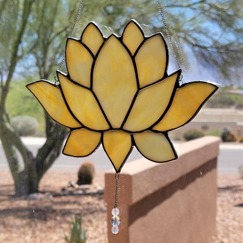 Peut inclure: Un attrape-soleil en forme de fleur de lotus en vitrail aux tons jaune et ambre. La fleur a des contours noirs et est suspendue par une cha&icirc;ne. Une petite cha&icirc;ne de perles pend du bas. L'arri&egrave;re-plan montre une sc&egrave;ne ext&eacute;rieure floue.