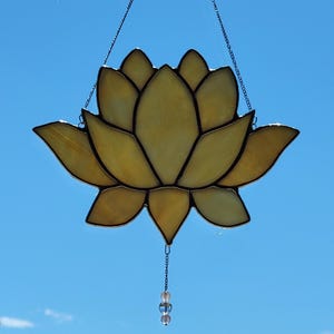 Peut inclure: Un vitrail en forme de fleur de lotus aux tons jaunes et ambr&eacute;s, avec des contours noirs. La fleur est suspendue par une cha&icirc;ne et poss&egrave;de un petit ornement de perles en bas. Le fond est un ciel bleu clair et lumineux.