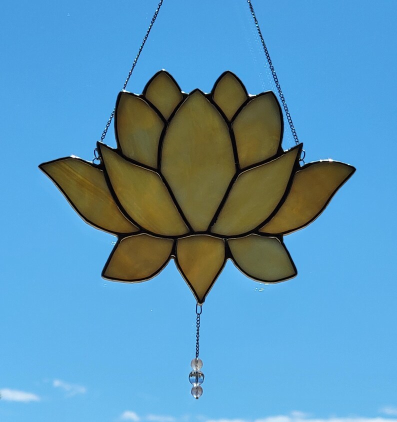 Peut inclure: Un vitrail en forme de fleur de lotus aux tons jaunes et ambr&eacute;s, avec des contours noirs. La fleur est suspendue par une cha&icirc;ne et poss&egrave;de un petit ornement de perles en bas. Le fond est un ciel bleu clair et lumineux.