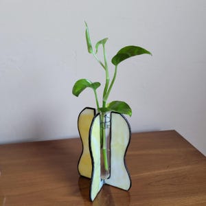 May include: A yellow and white stained glass vase with a black outline holds a single green plant in a glass test tube.