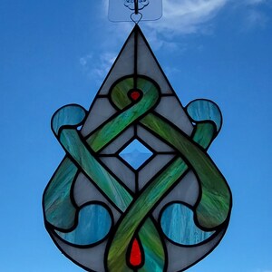 May include: A stained glass teardrop-shaped ornament with a green, blue, and white design. The ornament features a Celtic knot pattern with a diamond-shaped center.