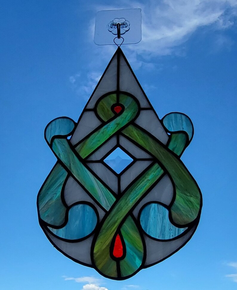 May include: A stained glass teardrop-shaped ornament with a green, blue, and white design. The ornament features a Celtic knot pattern with a diamond-shaped center.