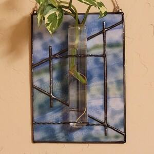May include: A rectangular stained glass plant holder with a clear glass tube for a plant cutting. The glass has a blue and white watercolor effect, framed by dark metal lines. A green and white plant cutting is visible.