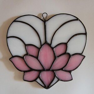 May include: Stained glass heart-shaped suncatcher featuring a pink lotus flower design. The lotus is surrounded by white glass, all framed with black solder. Includes a small metal loop for hanging. A decorative piece for windows or walls.