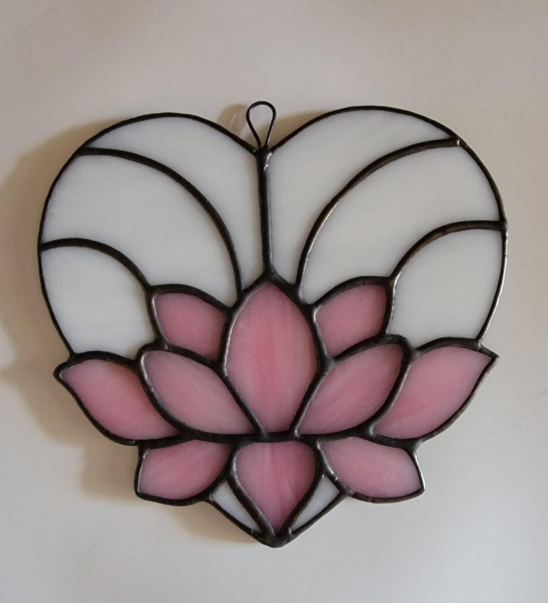 May include: Stained glass heart-shaped suncatcher featuring a pink lotus flower design. The lotus is surrounded by white glass, all framed with black solder. Includes a small metal loop for hanging. A decorative piece for windows or walls.
