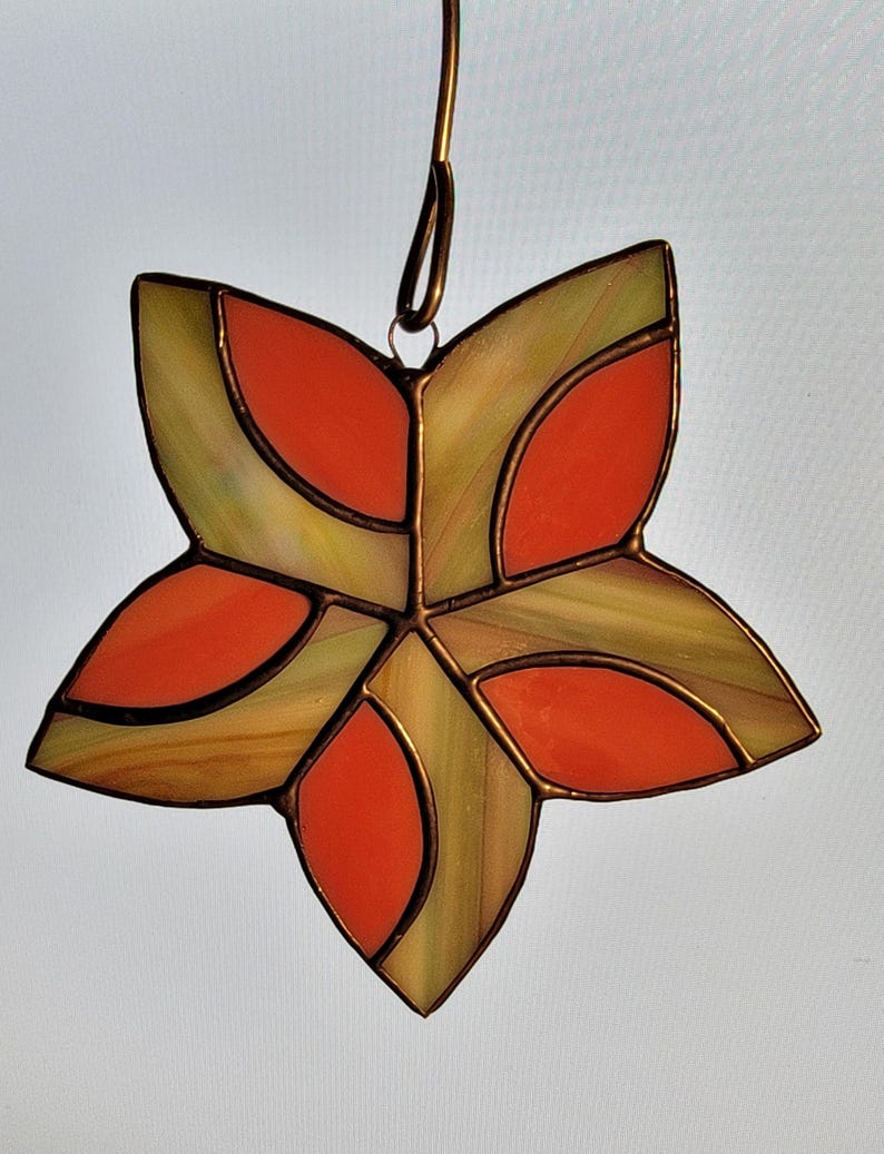 K&ouml;nnte beinhalten: Ein Ornament aus Buntglas in Form eines Sterns mit geometrischem Muster. Der Stern besteht aus orangem und gr&uuml;nem Glas und hat einen Aufh&auml;nger aus Kupferdraht.