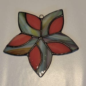 K&ouml;nnte beinhalten: Ein Ornament aus Buntglas in Form eines Sterns mit rotem und gr&uuml;nem Design. Der Stern besteht aus mehreren Glasst&uuml;cken, die mit Blei zusammengef&uuml;gt sind.