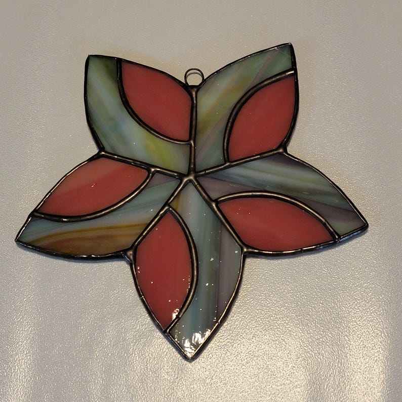 K&ouml;nnte beinhalten: Ein Ornament aus Buntglas in Form eines Sterns mit rotem und gr&uuml;nem Design. Der Stern besteht aus mehreren Glasst&uuml;cken, die mit Blei zusammengef&uuml;gt sind.