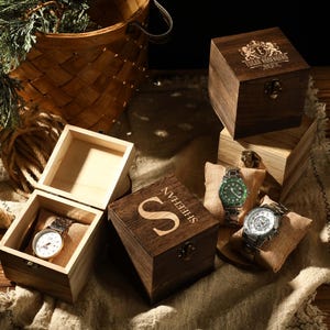 Może przedstawiać: Drewniane pudełka na zegarki z personalizowanymi grawerunkami, prezentujące srebrny i zielony zegarek. Zegarki są prezentowane na jutowych poduszkach. Jedno pudełko jest otwarte, ukazując zegarek z białą tarczą. W tle drewniany kosz.
