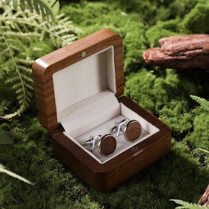 May include: A wooden box with a light-colored interior holds a pair of cufflinks. The cufflinks have a brown circular design with silver-colored metal accents. The box is open, revealing the cufflinks, and sits on a bed of green moss.