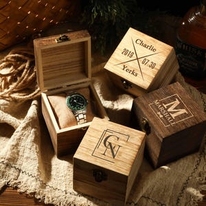 Może przedstawiać: Drewniane pudełka na zegarki z personalizowanymi grawerunkami. Jedno pudełko jest otwarte, ukazując zegarek z zieloną tarczą i srebrną bransoletą. Inne pudełka zawierają imiona "Charlie", "Marshall" i inicjały. Na lnianej tkaninie.