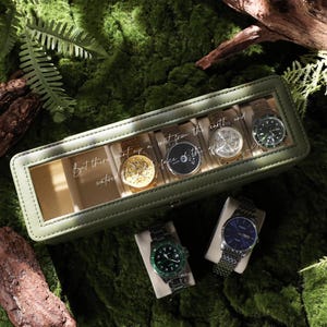 May include: An olive green watch case with a clear top, holding five watches. The case is surrounded by moss and greenery. Two additional watches are displayed below. The text on the case reads, "But there went up a watered face of the nature."