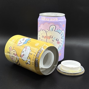 May include: Two decorative metal cans with cartoon illustrations. One is yellow with a bee and bunny design, the other is purple with a bear design. Both cans have the text "OCEAN BOMB". A white plastic cap is next to the yellow can.