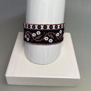 May include: A beaded bracelet with a black background and a floral pattern in white and brown. The bracelet is displayed on a white cylindrical stand. The floral design includes small, white, four-petal flowers and brown accents.