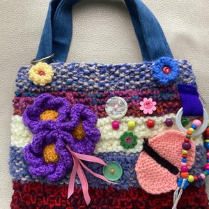 Puede incluir: Una bolsa de mano de punto de colores con una correa de mezclilla. La bolsa está decorada con flores de ganchillo, botones, cuentas y una mariposa rosa.